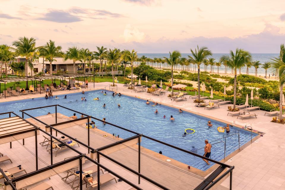 Hotel pool with many people swimming and relaxing on lounge chairs near the beach.