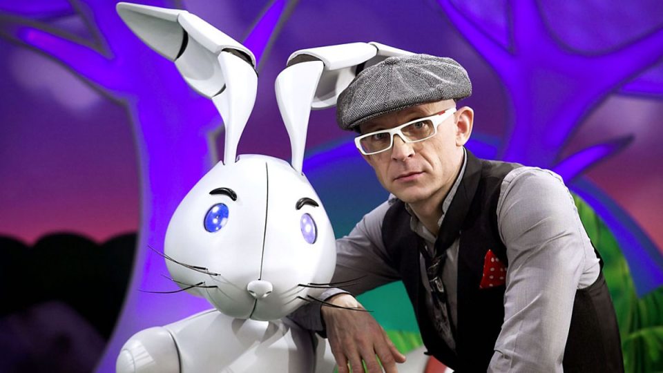 A man with a newsboy cap and glasses poses with a robotic rabbit.