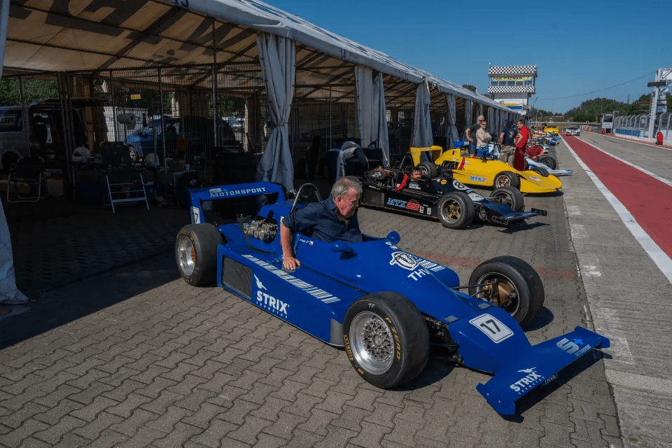 A man sits in a blue Formula One race car in a pit lane.