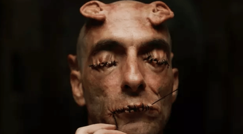 Close-up of a man with stitched eyes and mouth, and small horns on his head.