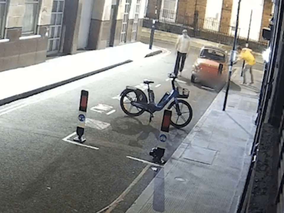 Screenshot of a parked bicycle near a red car and pedestrians on a city street.