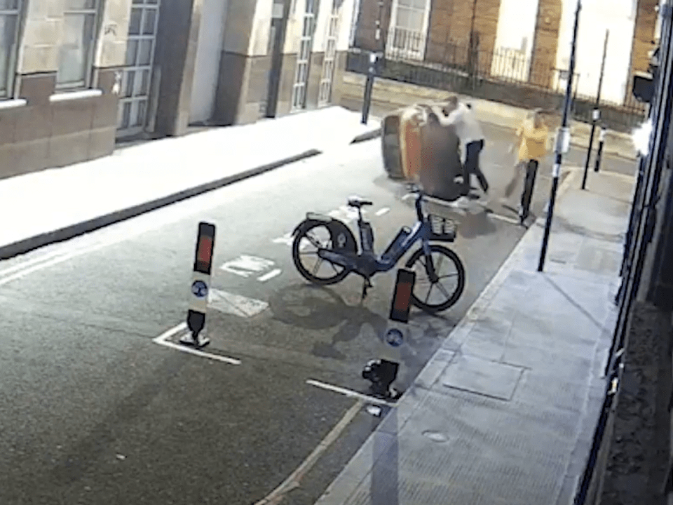 Screenshot of two people struggling with a large object on a city street, a bicycle is parked nearby.