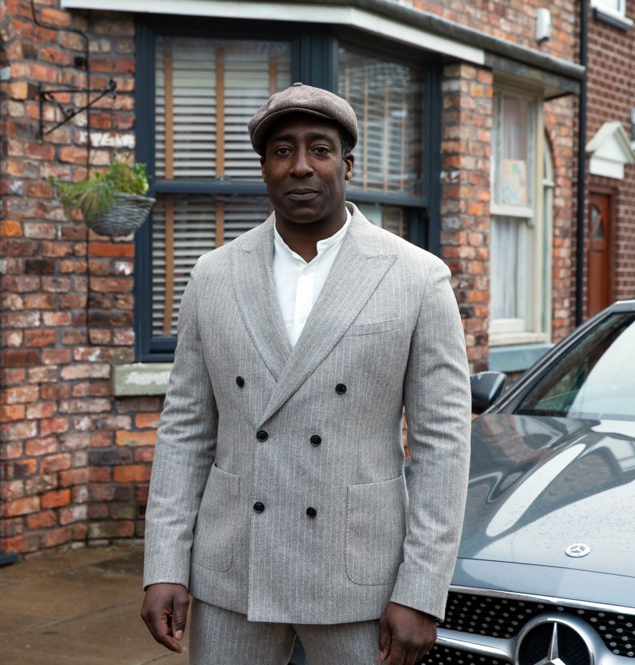 A man in a gray suit stands in front of a brick building and a sports car.