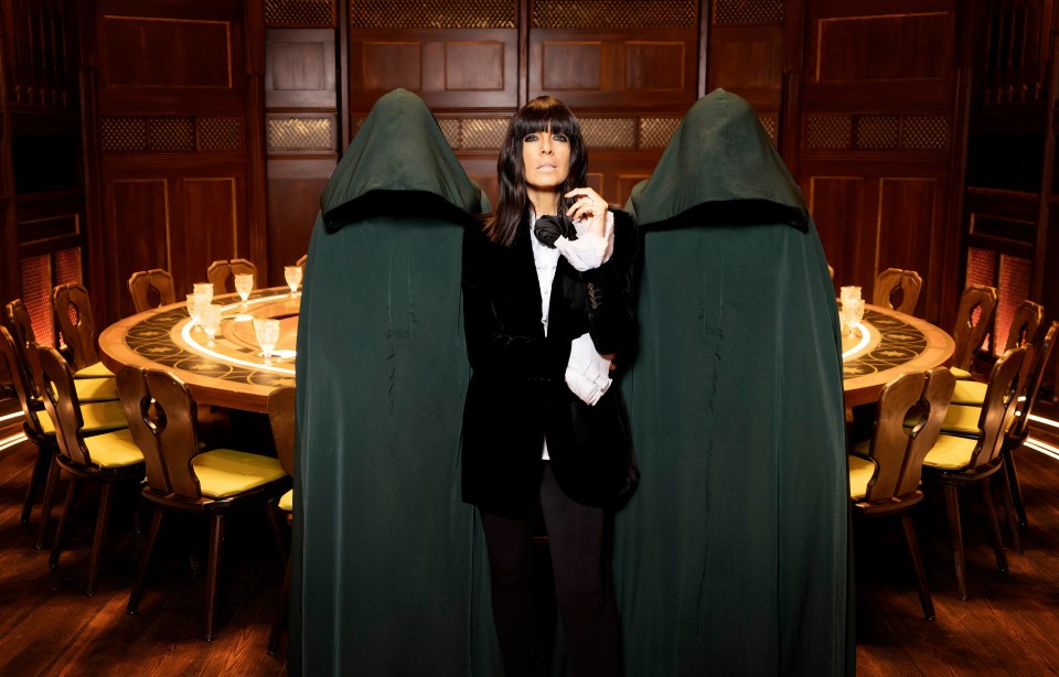 Claudia Winkleman on the set of The Traitors, flanked by two figures in hooded robes.