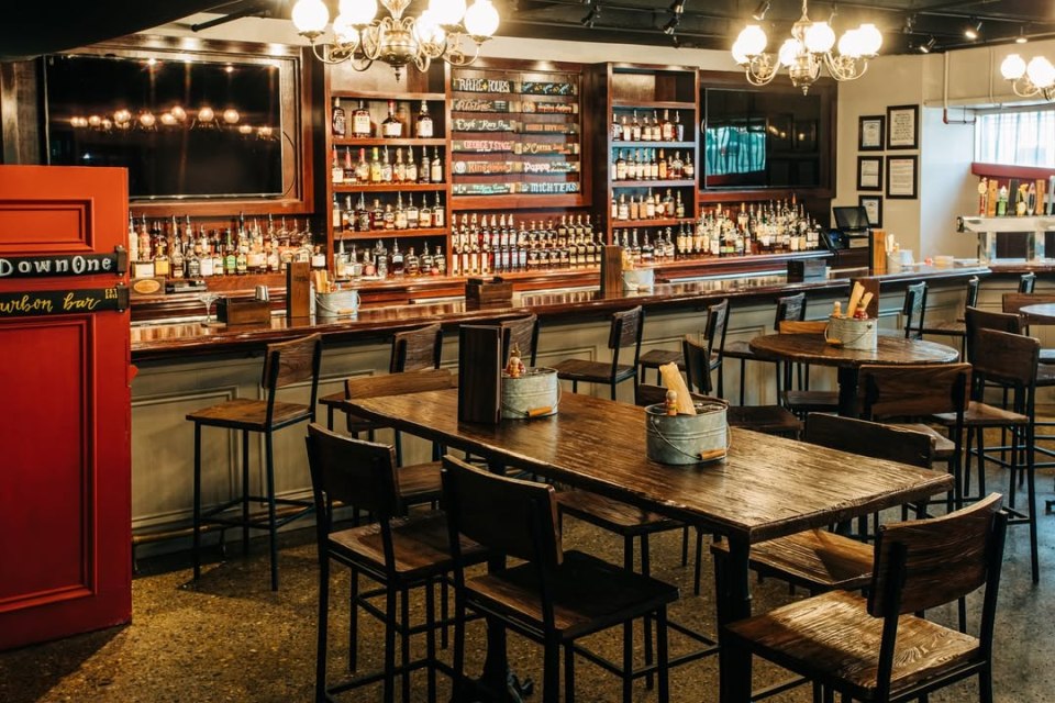 Interior view of Down One Bourbon Bar in Louisville, showing a long bar stocked with bourbon bottles and several wooden tables and chairs.