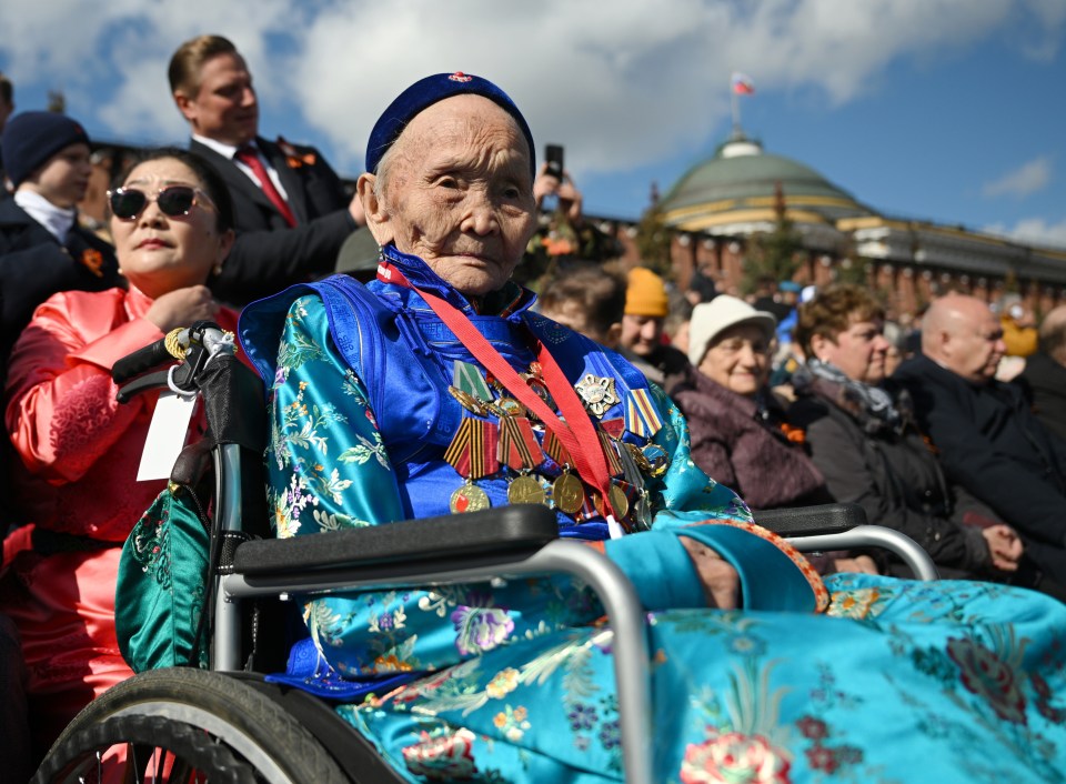 Elderly woman in wheelchair wearing medals at a military parade.