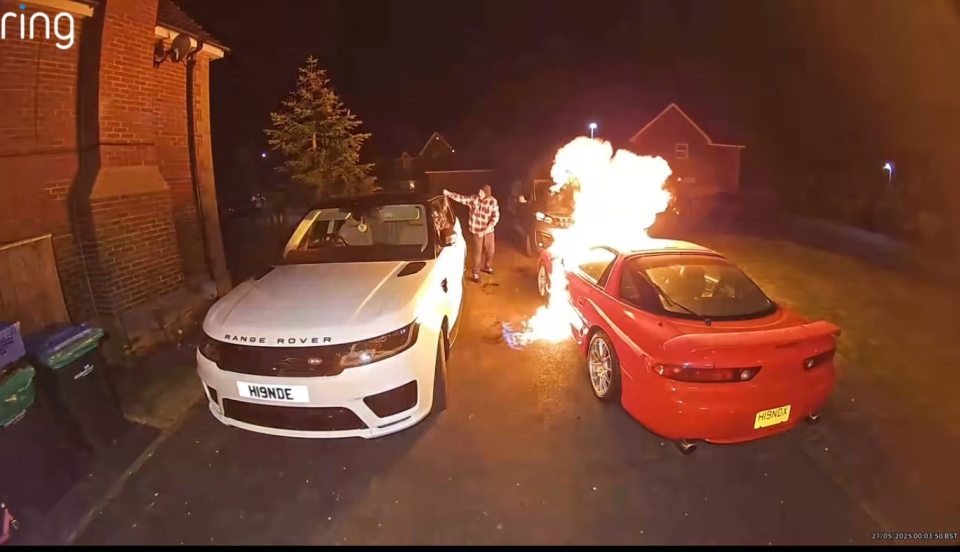 A man watches as a red car burns in a driveway next to a white Range Rover.