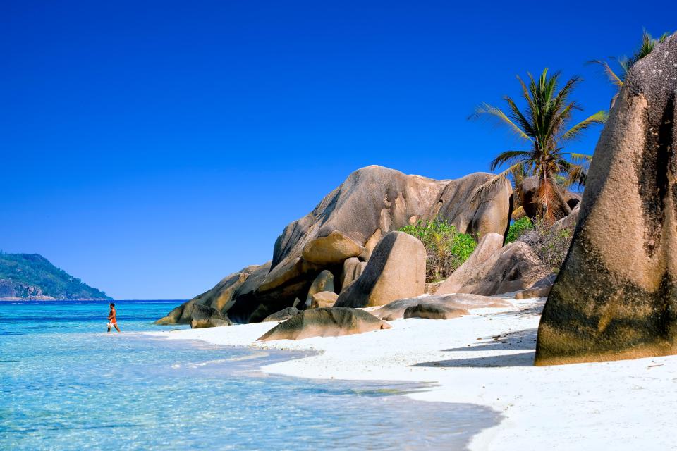 Person wading in shallow turquoise water on a white sand beach with large granite boulders and a palm tree.