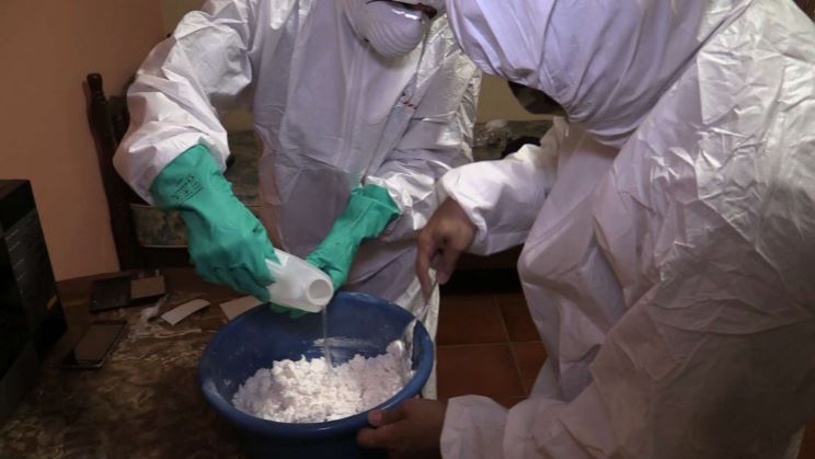 Two people in protective suits mixing a white powder.