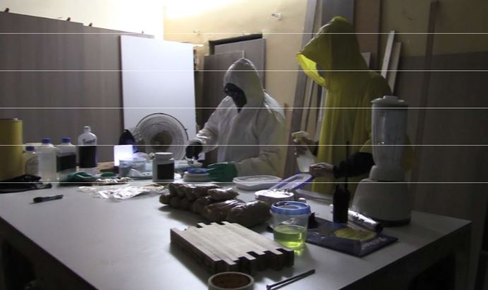 Two people in protective suits working with chemicals and a blender.