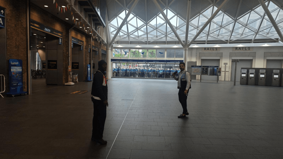 Two men standing in a train station with a large crowd visible through a glass wall.