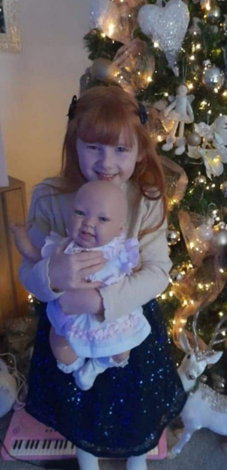 Girl holding a baby doll in front of a Christmas tree.