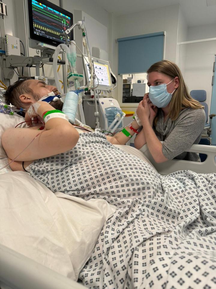 A woman in a face mask holds the hand of a man lying in a hospital bed.