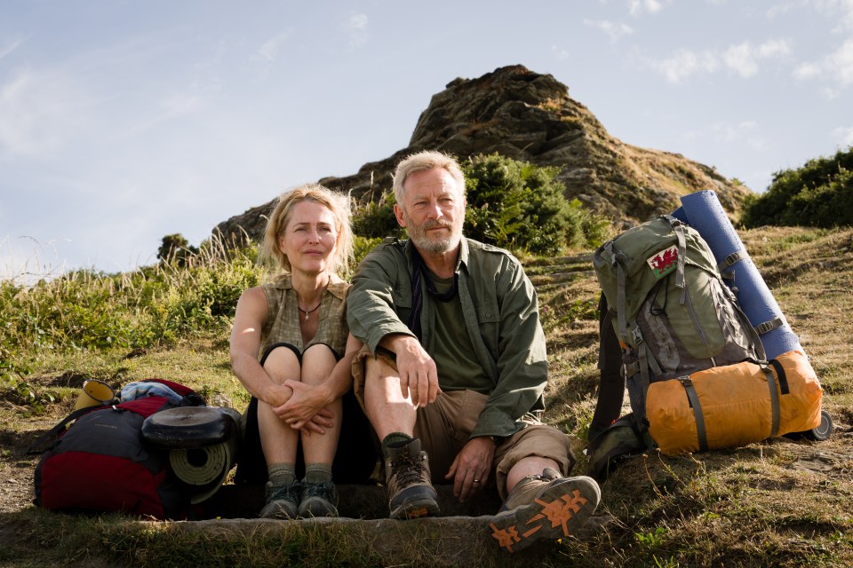 Gillian Anderson and Jason Isaacs sitting on a hill with backpacks.
