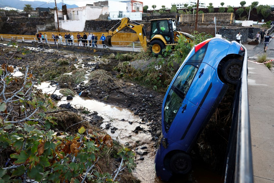 A blue car swept away by floodwaters rests against a railing; onlookers stand nearby.