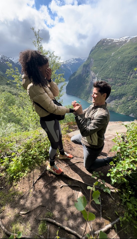 A man proposing to a woman on a mountain overlooking a fjord.