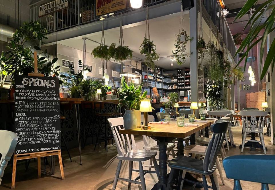 Interior view of a restaurant with hanging plants, a chalkboard menu, and tables set for diners.