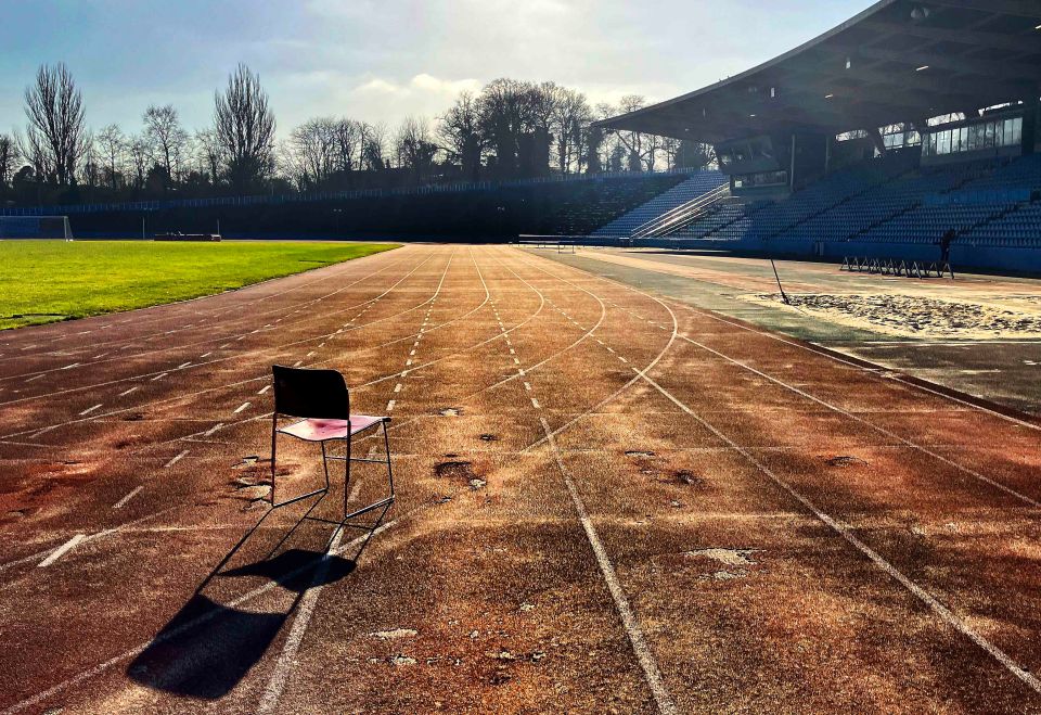 A single chair sits on a weathered running track in front of an empty stadium.