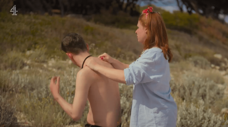 A woman applies sunscreen to a man's back on a beach.