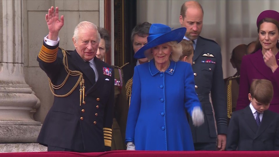 King Charles III and Camilla, Queen Consort, waving from a balcony.  Prince William and Catherine, Princess of Wales, are visible in the background.