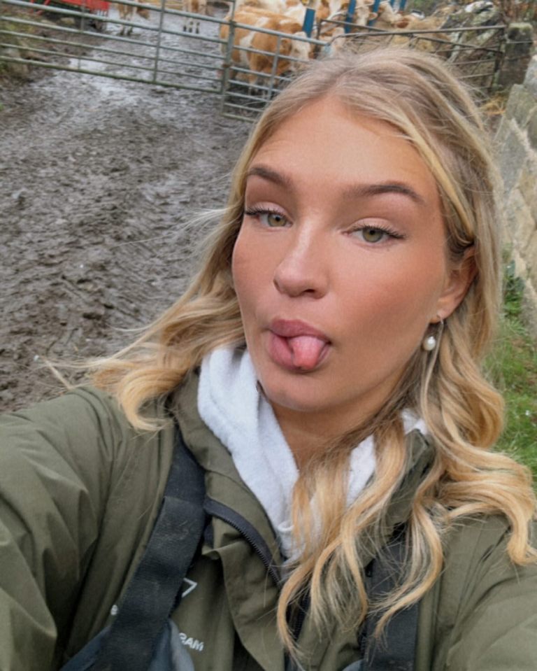 A blonde woman sticking her tongue out near cows in a muddy pen.