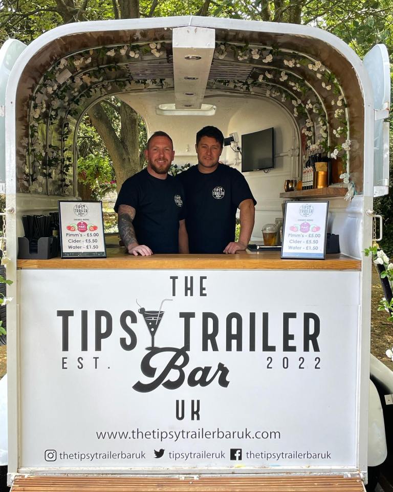 Two men standing behind the bar of a mobile bar called "The Tipsy Trailer Bar".