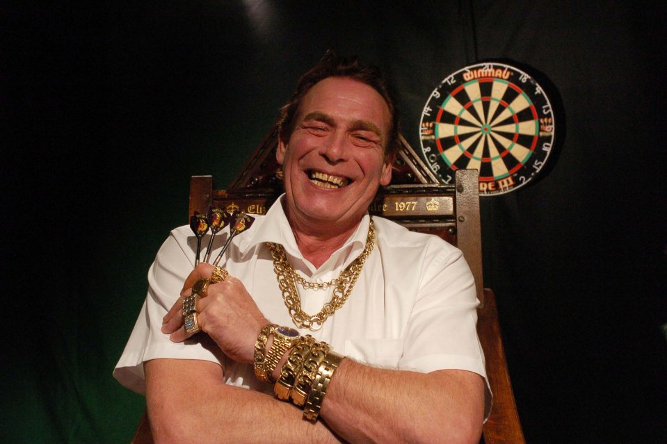 A smiling darts player wearing many gold chains and rings, holding darts.  A dartboard is in the background.