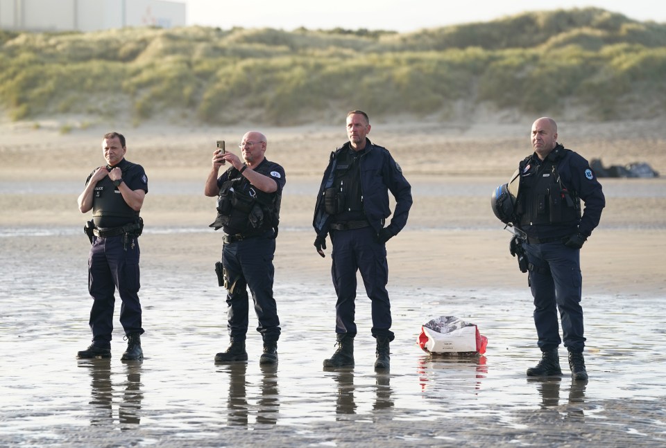 French police officers on a beach watching a group of people board a small boat.